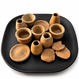 May include: A collection of small, unglazed terracotta pottery pieces including several miniature vessels, bowls, and flat circular discs. These earthy, light brown clay items are arranged together on a smooth, matte black square plate.