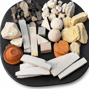 Puede incluir: Una colección de varias piezas de arcilla y tiza naturales dispuestas sobre un plato negro. El surtido incluye rocas blancas, grises, amarillas y anaranjadas, palitos rectangulares, esferas lisas y un pequeño cuenco circular de color terracota.
