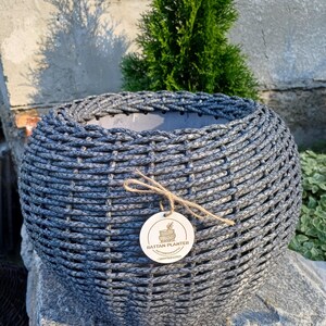 Puede incluir: Una maceta redonda de ratán trenzado de color gris oscuro con un exterior texturizado. Una pequeña etiqueta redonda de madera está atada al frente con cordel, con el texto RATTAN PLANTER, un dibujo lineal de una maceta y un número de teléfono.