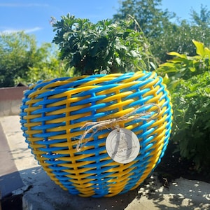 Puede incluir: Una maceta redonda de ratán trenzado con un vibrante patrón alterno de hebras de color azul brillante y amarillo. Una pequeña etiqueta blanca circular está atada al frente con cordel, mostrando el texto RATTAN PLANTER y datos de contacto.