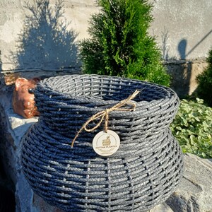 Könnte beinhalten: Ein dunkelgrauer, geflochtener Rattan-Pflanzkübel mit bauchiger Form und weiter Öffnung. Ein dünnes braunes Band ist um den Rand gebunden und hält ein kleines, rundes Holzschild mit einer Illustration und dem Text RATTAN PLANTER.