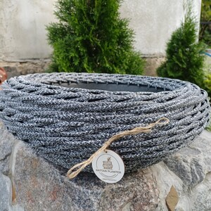 Può includere: Un vaso rotondo a profilo basso con un esterno intrecciato in materiale sintetico maculato bianco e nero. Un'etichetta rotonda in legno pende dal lato tramite un sottile spago di juta, riportando il testo RATTAN PLANTER e un numero di telefono.