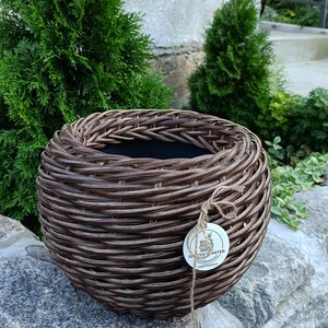 May include: A round, dark brown woven rattan planter with a wide, braided rim and a deep, hollow center. A small, circular white tag with the text RATAN PLANTER is attached to the side of the planter with a thin, light brown twine string.