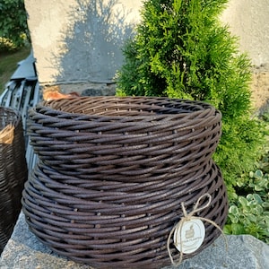 Puede incluir: Una maceta redonda de ratán marrón oscuro con textura tejida y un borde ancho y acampanado. Una pequeña etiqueta blanca circular está atada al lateral con cordel, mostrando un gráfico de una cesta y el texto RATTAN PLANTER en tinta negra.