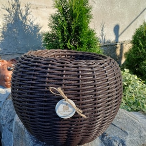 Könnte beinhalten: Ein runder, dunkelbrauner geflochtener Rattan-Pflanzkübel mit einer breiten, offenen Oberseite. Ein kleines, rundes weißes Etikett ist mit einer Schnur an der Vorderseite befestigt, mit einer Zeichnung des Pflanzkübels und dem Text RATTAN PLANTER.