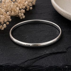 以下が含まれることがあります： 丸みを帯びたプロファイルの滑らかで磨き上げられたシルバーのバングルブレスレット。円形のバンドは光を捉える反射性の金属表面を備えており、継ぎ目のない連続した頑丈な金属構造に沿って繊細なハイライトを作り出しています。
