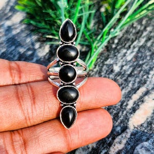 May include: A silver-toned ring featuring five vertically aligned, smooth black stones. The design includes two teardrop-shaped stones at the top and bottom, with three oval-shaped stones positioned in the center, all set within a metallic band.