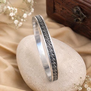 May include: A silver-toned metal bangle bracelet featuring a continuous, raised, dark-oxidized rope or twisted cable pattern running along the center of the outer band, with smooth, polished silver edges on both sides of the textured design.