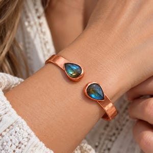 May include: A copper cuff bracelet featuring two teardrop-shaped labradorite gemstones at the open ends. The metal band has a smooth, polished finish, and the stones display iridescent blue and green flashes against the warm metallic surface.