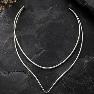 May include: A silver-toned metal choker necklace featuring a double-strand design. The upper strand is a smooth, rounded arc, while the lower strand forms a distinct V-shape point at the center. The necklace has an open back construction.