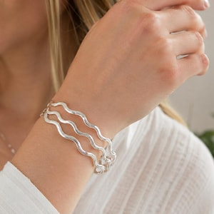 May include: A set of three silver-toned bangles, each featuring a consistent wavy or serpentine design. The bracelets have a smooth, reflective metallic finish and are worn stacked together on the wrist, creating a layered appearance.