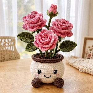 Könnte beinhalten: Eine gehäkelte Topfpflanze mit drei blühenden rosa Rosen und einer Rosenknospe mit grünen Blättern. Der runde, cremefarbene Topf hat einen dunkelbraunen Rand und Boden sowie ein gesticktes lächelndes Gesicht mit schwarzen Augen und rosa Wangen.