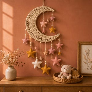 Può includere: Una decorazione da parete composta da una grande mezzaluna in macramè color crema, ornata da numerose stelle in feltro pendenti nei toni del rosa, giallo senape e bianco, sospese tramite fili con piccole perline di legno inserite.