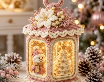 May include: A decorative crochet lantern in dusty pink featuring a scalloped trim, a top handle, and a floral accent. Inside, a crocheted snowman wearing a pink hat and scarf stands next to a white crocheted Christmas tree with gold beads.