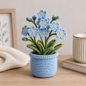 May include: A crocheted potted plant featuring light blue flowers with small yellow centers and green leaves. The flowers are arranged on thin green stems emerging from a textured, light blue crocheted pot with a rounded, cylindrical shape.