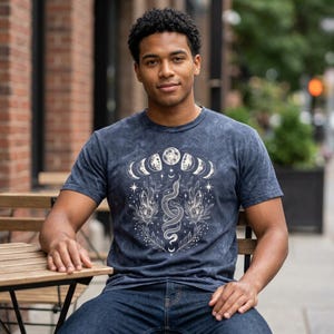 以下が含まれることがあります： ダークブルーのアシッドウォッシュTシャツ。胸元には、クリスタルと植物が絡み合う中央の蛇と、その周囲を半円状に囲む月相と小さな星々が描かれた白いグラフィックプリントが施されています。