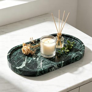 May include: An oval dark green marble tray with white veining. The tray holds a clear glass candle holder with a white wax candle, a small glass reed diffuser with four wooden reeds, a dried beige flower, and a small green leafy branch.