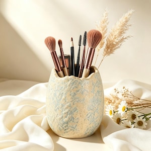 Puede incluir: Un soporte de cerámica en forma de huevo con un acabado texturizado moteado en crema y azul claro, que contiene varios pinceles de maquillaje, un lápiz y una pequeña paleta de sombras, decorado con hierba de la pampa y margaritas blancas.