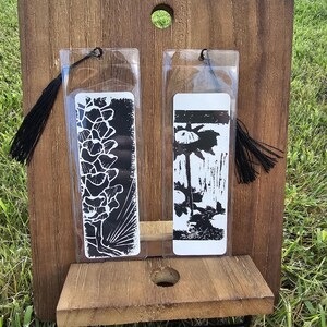 Può includere: Due segnalibri rettangolari in custodie di plastica trasparente, ciascuno con una stampa botanica in bianco e nero. Il segnalibro a sinistra mostra un motivo floreale, quello a destra un girasole. Entrambi hanno nappe nere attaccate in alto.