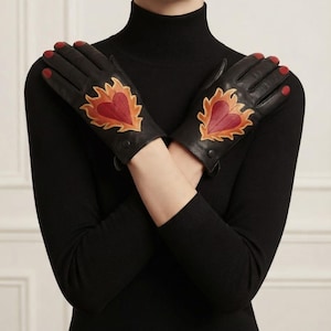 Peut inclure: Une paire de gants en cuir noir arborant une application en forme de cœur rouge entourée de détails en forme de flammes orange et jaunes sur le dos de chaque main. Les gants présentent une finition lisse et sont portés les bras croisés.
