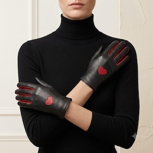 Könnte beinhalten: Ein Paar schwarze Lederhandschuhe mit roten Lederakzenten an den Fingerzwischenräumen und einem kleinen roten herzförmigen Aufnäher auf dem Handrücken. Die Handschuhe haben eine glatte Oberfläche und bedecken die Hände und Handgelenke.