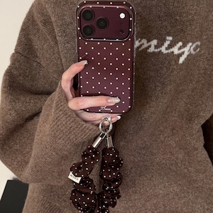 May include: A dark maroon smartphone case featuring a white polka dot pattern and a matching dark maroon fabric wrist strap with white polka dots. The case has a camera cutout and the text T Fanas printed at the bottom, with a small white tag on the strap.
