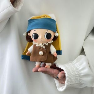 May include: A small doll with large black eyes, wearing a brown vest over a white shirt with a lace collar, and a blue and yellow headpiece featuring two white pearl-like accents on the sides and a single pearl button on the front of the vest.