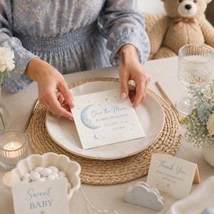 Puede incluir: Una servilleta de papel cuadrada blanca con una ilustración de una luna creciente azul con cara, pequeñas estrellas doradas y el texto "Over the Moon For Baby Oliver James Sweet Little Dreams 11.03.2027" en letras azules y doradas.
