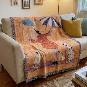 May include: A woven throw blanket featuring an orange background with a person holding a black and white umbrella, a blue umbrella, a dog, and a goose. The blanket has fringed edges and displays a whimsical illustration of a figure in a coat.
