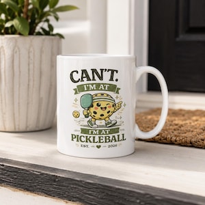 May include: A white ceramic mug featuring a cartoon pickleball character holding a paddle. The text reads: CAN'T. I'M AT I'M AT PICKLEBALL EST. 2026 with a small heart icon. The design uses dark green and yellow colors with star accents.
