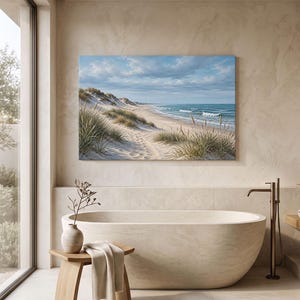 May include: A rectangular wall art print depicting a coastal landscape with sand dunes, tall beach grass, and a blue ocean under a cloudy sky. The scene is displayed above a smooth, oval-shaped beige bathtub and a small wooden stool with a towel.