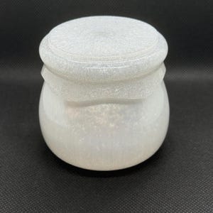 May include: A small, round, white jar with a matching lid, featuring a glossy, pearlescent finish with subtle glitter particles throughout the material. The container has a smooth, curved body and a slightly raised, tiered lid design.