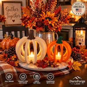 May include: Two hollow pumpkin-shaped candle holders, one white and one orange, sit on a marble tray. Each holds a small lit tea light. They are surrounded by autumn-themed decor, including a metal pitcher with sunflowers, berries, and fall leaves.