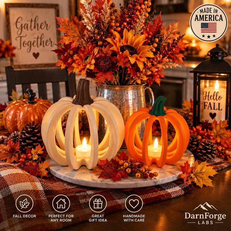 May include: Two hollow pumpkin-shaped candle holders, one white and one orange, sit on a marble tray. Each holds a small lit tea light. They are surrounded by autumn-themed decor, including a metal pitcher with sunflowers, berries, and fall leaves.