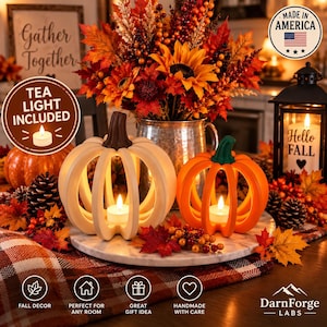 May include: Two decorative pumpkin-shaped candle holders, one cream and one orange, each featuring a tea light inside. They are arranged on a marble tray surrounded by autumn-themed foliage, pinecones, and a black lantern with a Hello Fall sign.