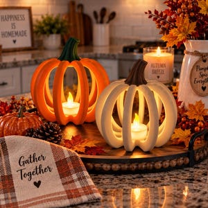 May include: Two open-frame pumpkin candle holders, one orange and one white, each containing a small LED tea light. They sit on a dark wooden tray with a plaid cloth featuring the text Gather Together, a small decorative pumpkin, and a white vase.