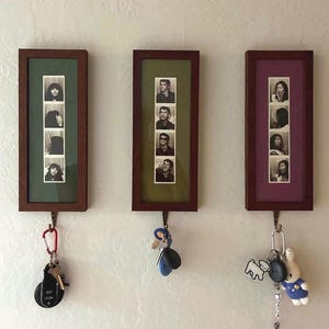May include: Three rectangular wooden wall-mounted key holders, each featuring a vertical black and white photo strip matted in green, olive, and purple. Each frame includes a metal hook at the bottom holding various keys, keychains, and charms.
