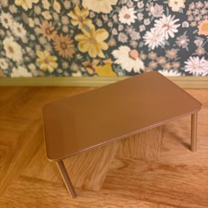 May include: A small, rectangular, light brown table with a smooth, flat top surface and four thin, straight legs. The table is designed with rounded corners and a solid, uniform color finish, presenting a simple, functional structure for display.