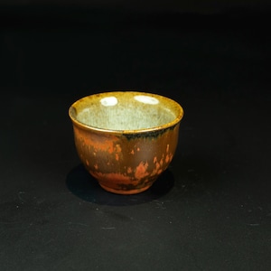 May include: A small ceramic cup featuring a mottled exterior with shades of deep brown, burnt orange, and black. The interior of the cup has a light, pale grey glaze that contrasts with the dark, earthy tones of the outer surface and rim.