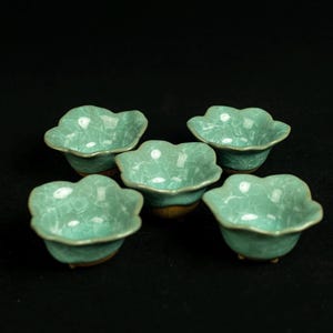 May include: A set of five small ceramic bowls with a light turquoise glaze and a distinct crackle pattern. Each bowl features a scalloped, flower-like rim and a brown, unglazed base, arranged in a cluster against a dark, solid background.