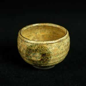 May include: A small, round ceramic bowl featuring a speckled glaze in shades of tan, beige, and dark green. The exterior surface has a subtle ribbed texture, and the rim is smooth and rounded, showcasing a variegated, earthy color palette.