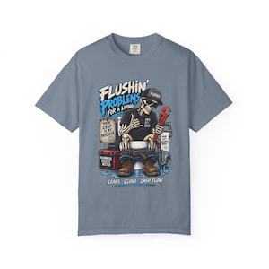 Op de afbeelding: Een leiblauw t-shirt met een afbeelding van een skelet-loodgieter die op een toilet zit en een rode pijptang vasthoudt. De tekst luidt: FLUSHIN' PROBLEMS FOR A LIVING, YOUR CRAP IS MY PAYCHECK, PIPE IT GOOD, PLUMBERS MAKE IT BETTER, LEAKS CLOGS CASH FLOW.