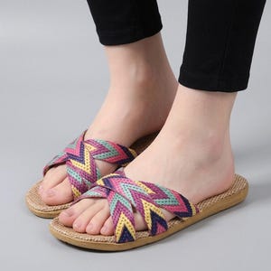 Puede incluir: Un par de sandalias sin cordones con tiras cruzadas con un patrón de chevrón tejido en tonos morado, rosa, amarillo y verde azulado. La plantilla tiene un acabado de material tejido de color tostado con textura, base plana para el calzado.