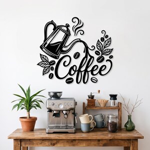 Könnte beinhalten: Ein schwarzes Wandkunstobjekt aus Metall, das eine stilisierte French Press zeigt, die Flüssigkeit in den in Schreibschrift geschriebenen Schriftzug Coffee gießt, umgeben von Kaffeebohnen und Blättern. Montiert an einer weißen Wand.