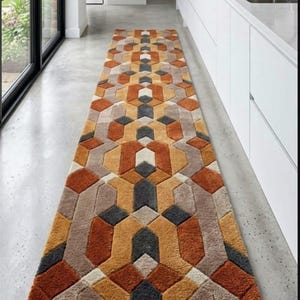 May include: A long rectangular runner rug featuring a repeating geometric pattern in shades of burnt orange, mustard yellow, charcoal grey, and cream. The design consists of interlocking hexagonal and arrow-like shapes with a textured pile surface.