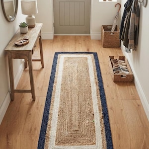 Puede incluir: Una alfombra de pasillo rectangular con un centro trenzado de color tostado natural, un borde blanco y un contorno exterior azul marino oscuro. La alfombra está hecha de un material de fibra texturizada y colocada sobre un suelo de madera.