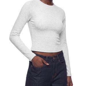 May include: A light grey heathered long-sleeve crew neck top with a fitted silhouette and a cropped hemline, paired with dark indigo denim jeans featuring a classic button fly and front pockets, creating a simple and neutral ensemble.