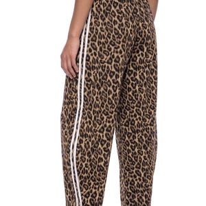 May include: Leopard print sweatpants featuring a brown and black animal pattern with two vertical white stripes running down the outer side of each leg. The pants have an elasticized waistband and a relaxed fit, extending to the lower calf area.