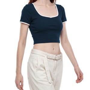 May include: A dark navy blue short-sleeved crop top featuring a white trim along the scoop neckline and sleeve cuffs. The fitted garment has a sweetheart-style neckline detail and is paired with high-waisted off-white trousers with a belt.