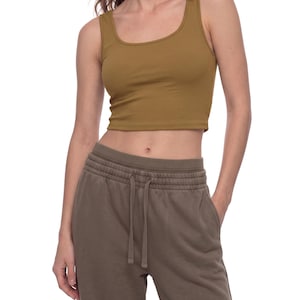 Może przedstawiać: Musztardowy, bezrękawnik typu crop top z okrągłym dekoltem w połączeniu z oliwkowymi spodniami dresowymi z elastycznym pasem i sznurkiem. Top jest dopasowany, natomiast spodnie dresowe mają luźny krój z widocznymi kieszeniami po bokach.